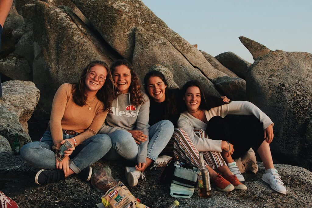 Group of cheerful young female friends in casual clothes laughing and looking at camera while sitting with drinks and snacks on rocky seashore and having fun during travel together