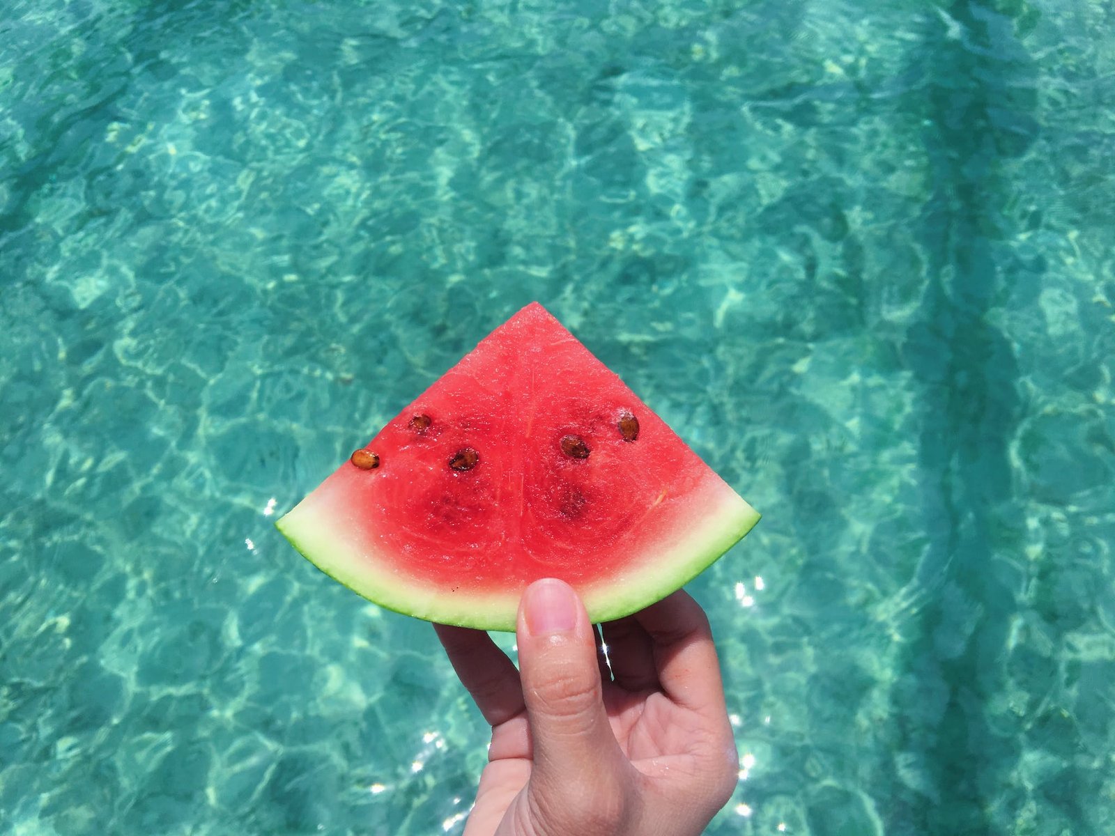 Hand Holding a Slice of Watermelon With Blue Swimming Pool Water in the Background on a hot day